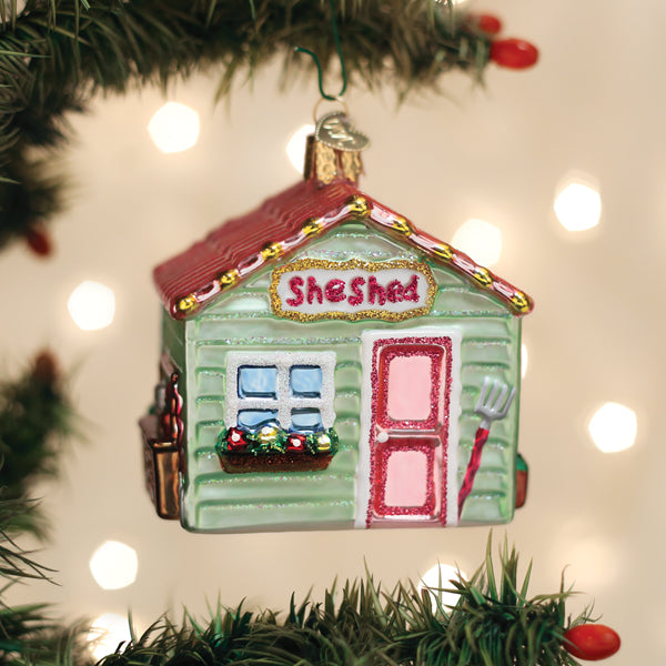 Decorative ornament shaped like a small house with 'She Shed' text, hanging on a Christmas tree.