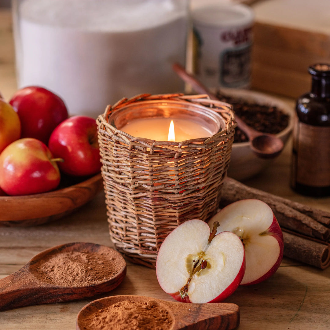 Candle in a woven holder with apples, cinnamon, and other spices on a wooden surface