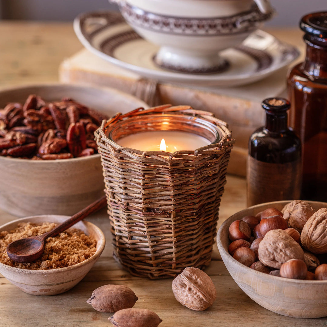 Wicker candle holder with a lit candle on a wooden table with bowls of nuts and spices.
